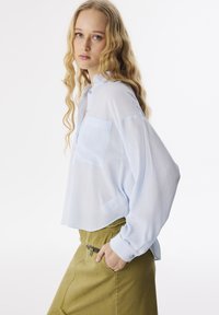 Mujer joven con cabello rubio largo y ondulado, vistiendo una blusa holgada de color azul claro y una falda verde oliva, con una mano en el bolsillo de la falda, de pie de lado.