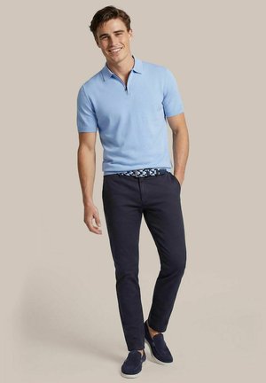 Junger Mann lächelnd, trägt hellblaues kurzärmeliges Poloshirt, dunkle enge Hose, gemusterten Gürtel und marineblaue Slipper, steht vor schlichten Hintergrund.