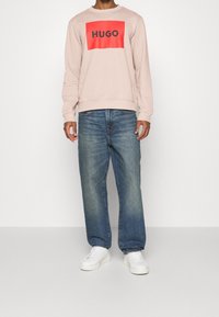 Hombre vistiendo un sudadera rosa claro con el logo rojo de "HUGO", jeans azules holgados y zapatillas blancas, de pie contra un fondo blanco liso.