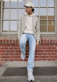 Beige checked blazer over a cream hoodie, light blue jeans, and gray sneakers. Setting against a brick wall with a window.