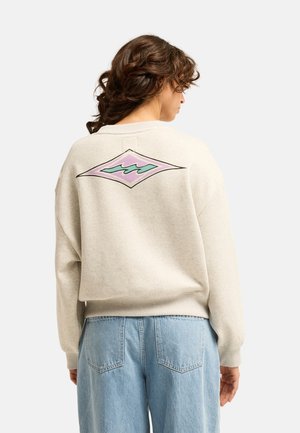 Vrouw met krullend haar die een beige sweatshirt draagt met een paarse en blauwgroene golf in diamantvorm op de rug en lichtblauwe spijkerbroek.