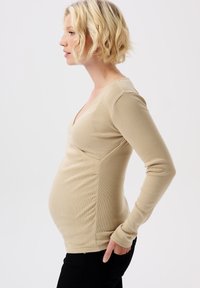 Hellbeige langärmliges Stilloberteil mit tiefem V-Ausschnitt, gerippter Textur, taillierter Passform und seitlicher Raffung zur Anpassung an einen wachsenden Bauch.