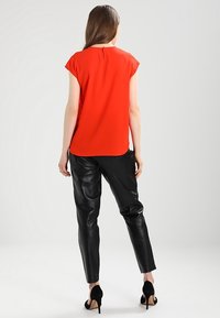 Mujer de pie con la espalda vuelta, vistiendo una blusa roja de mangas cortas, pantalones de cuero negros y zapatos de tacón negro sobre un fondo liso.