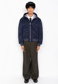 Marineblå quiltet bomberjakke med glidelås, ribbestrikkede mansjetter og kant. Brukt over en hvit skjorte og svart slips, kombinert med vide, grå bukser.