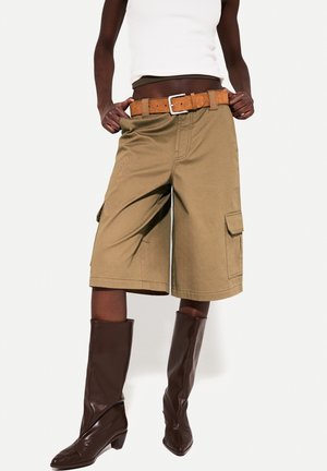 Person wearing knee-length beige cargo shorts with a brown belt, white tank top, and dark brown knee-high boots on a white background.