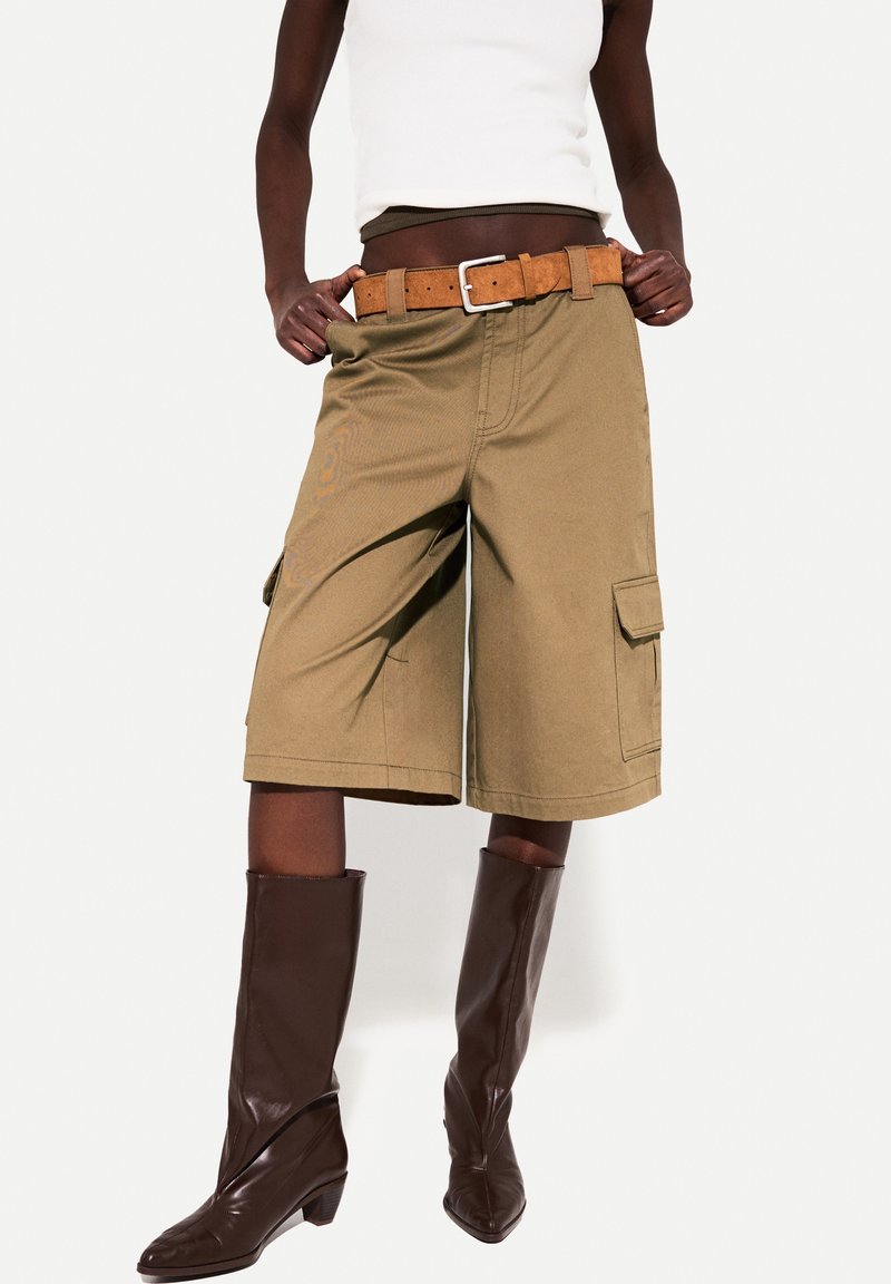 Personne portant un short cargo beige jusqu'aux genoux avec une ceinture marron, un débardeur blanc et des bottes hautes marron foncé sur fond blanc.