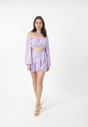 Ensemble deux pièces lilas comprenant un crop top à épaules dénudées avec des manches longues et une jupe portefeuille avec un ourlet à volants. Texture de tissu lisse.