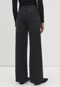 Un pantalon en denim noir taille haute, à jambe large, avec une texture lisse, doté de deux poches arrière et d'une coupe droite jusqu'aux chevilles.