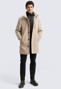 Beige mantel van een wolmix met een kraag, knoopsluiting en zijzakken, gedragen over een zwarte donsjas, gecombineerd met zwarte broek en laarzen.