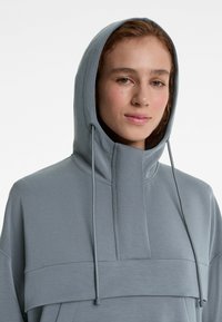 Sweatshirt cu glugă de culoare gri deschis, dotat cu un guler înalt, material texturat și sfori vizibile. Designul pune accent pe un aspect stratificat.
