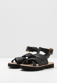 Schwarze gewebte Sandalen mit Riemen, die über ein strukturiertes Design und eine rechteckige Schnalle verfügen. Die Sohle ist schwarz mit einem Fußbett aus natürlichem Leder.