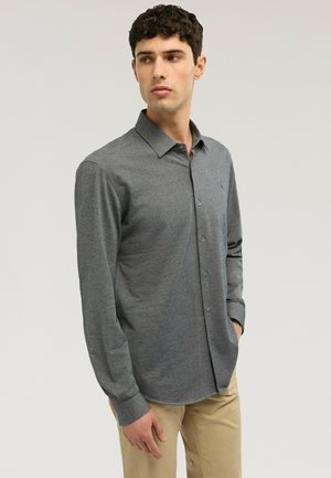 Uomo che indossa una camicia grigia a maniche lunghe con bottoni e pantaloni beige, guarda verso sinistra, con una mano in tasca.