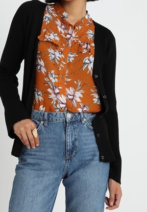 Schwarzer gerippter Cardigan über einer orangefarbenen geblümten Bluse mit Rüschen, kombiniert mit hoch taillierten hellblauen Jeans. Auffälliger Goldring an der Hand.