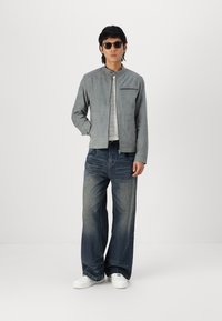 Jachetă din suede gri cu guler înalt și buzunar cu fermoar frontal, asortată cu blugi blue cu croi larg și adidași albi, model purtând ochelari de soare.