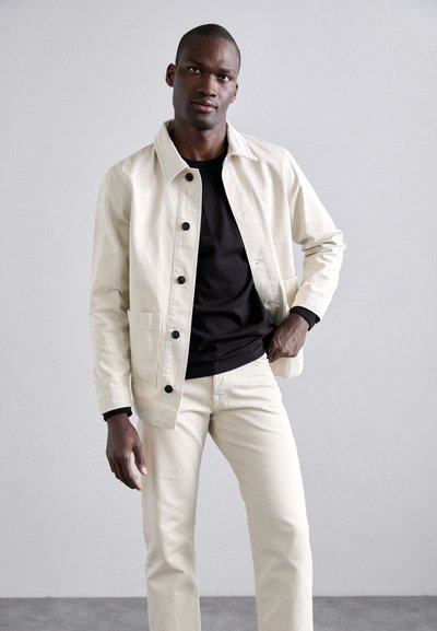 Light beige canvas jacket with dark buttons, black t-shirt, and light beige pants; smooth texture and tailored fit. Standing pose against a neutral background.