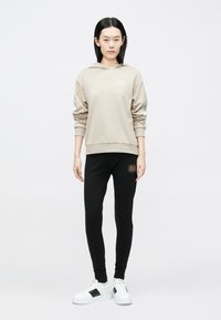 Bēša kapuci džemperis ar riboto aproci, kombinēts ar melnām legingām ar logotipu. Modelis valkā baltus sporta apavus ar melnām detaļām.