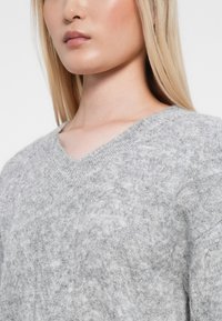 Harmaa neulottu villapaita, jossa on V-pääntie ja pehmeä, tekstuuri pinta. Kangas näyttää pörröiseltä hienovaraisella kuviolla.