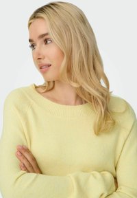 Pull en maille jaune clair avec un col large et des poignets côtelés. Texture douce et coupe décontractée, présenté avec les bras croisés.