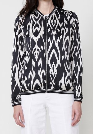 Vrouw draagt een zwart-wit patroon ritsjack met gestreepte manchetten en kraag, gecombineerd met een witte broek.