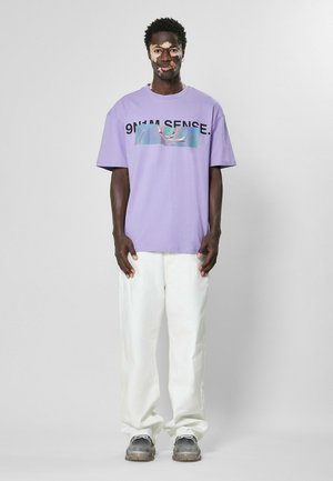 Lilla oversized t-skjorte med grafisk trykk og tekst, sammen med hvite bukser og grå sneakers på en person som står mot en lys bakgrunn.