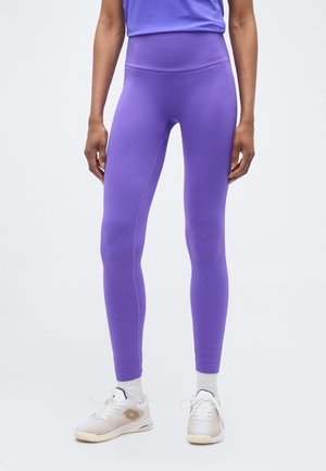 Άτομο που φοράει μωβ κολάν με ρίγες και ψηλή μέση, και λευκά αθλητικά παπούτσια με λευκές κάλτσες, στέκεται μπροστά σε απλό φωτεινό φόντο.