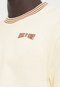 Κοντινό πλάνο ενός ατόμου που φοράει μια κρεμ φούτερ με καφέ διπλή ρίγα στον γιακά και το κέντημα «HOUSE OF SUNNY» στο στήθος.