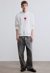 Λευκό φούτερ με κουκούλα, διακοσμημένο με κόκκινη καρδιά και το κείμενο «HUGO», σε συνδυασμό με φαρδιά γκρι τζιν και μαύρα παπούτσια. Λείος υφής.