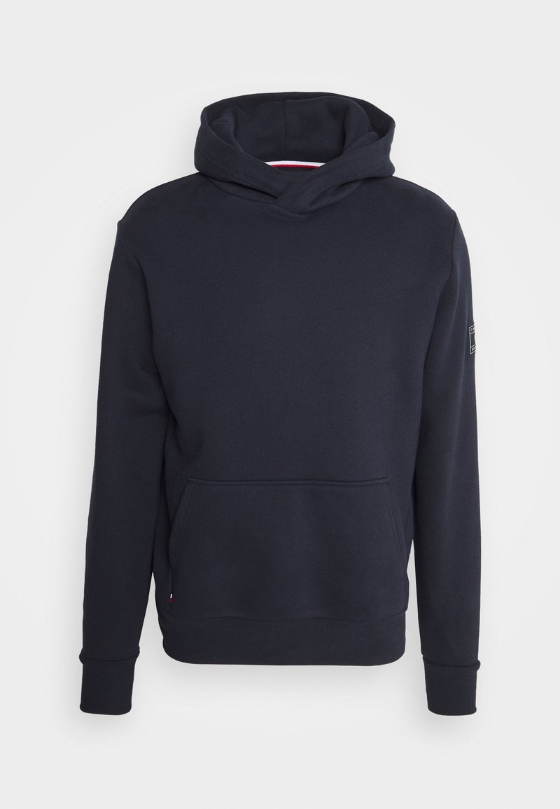 Sweat à capuche bleu marine foncé à manches longues, poche kangourou à l'avant et écusson sur le haut du bras gauche, présenté sur un fond uni.