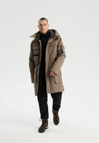 Długa, beżowa parka z kapturem, poziome czarne paski, boczne kieszenie i kieszeń na suwak na rękawie; noszona na czarnych spodniach i butach.