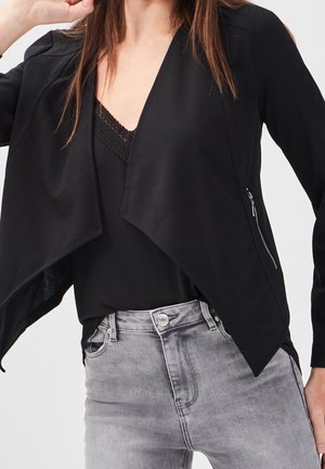 Femme portant une veste noire ouverte avec un détail de fermeture éclair sur un haut noir bordé de dentelle et un jean gris taille haute.