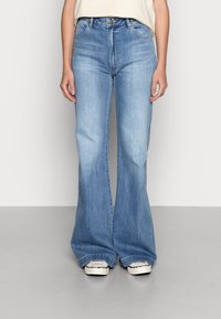 Jeans acampanados de color azul claro, hechos de denim, con cintura alta, un ligero desgaste y un diseño clásico de cinco bolsillos, combinados con zapatillas blancas.