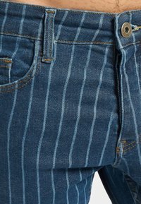 Sinised teksapüksid heledate vertikaalsete triipudega, nähtav nööp, vööaas, esi tasku ja õmbluse detailid.