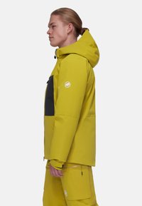 Gelbe wasserdichte Skijacke mit einer dunkelgrauen Brusttasche, Reißverschlüssen an den Bündchen und einem Logopatch am Ärmel. Verstellbare Kapuze.
