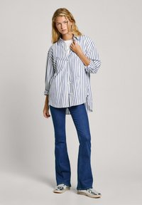 Camicia a righe blu e bianca, maniche lunghe, con bottoni davanti, abbinata a jeans a zampa d'elefante. Il modello si presenta in modo informale, con le mani sulla camicia.