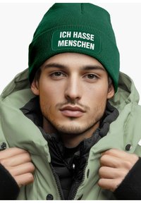 Green knitted beanie with a patch reading "ICH HASSE MENSCHEN." The subject wears a light green and black jacket, showcasing a layered look.