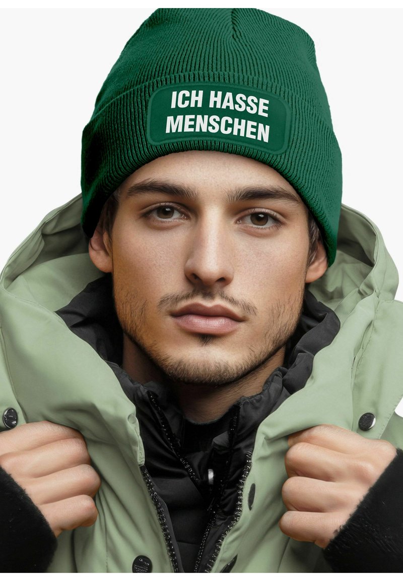 Green knitted beanie with a patch reading "ICH HASSE MENSCHEN." The subject wears a light green and black jacket, showcasing a layered look.
