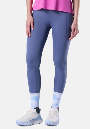 Femme portant des leggings bleus ajustés, un haut rose, des chaussettes blanches à motifs et des chaussures de sport blanches, debout devant un fond uni.