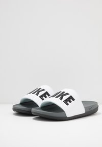 Baltos šlepetės su plačiu dirželiu, ant kurio ryškiai juodai parašyta "NIKE". Pilka pado spalva, lygi, matinė apdaila.