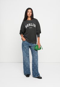 Μαύρη φούτερ μπλούζα με λευκό κείμενο "BERLIN", μπλε τζιν με μοτίβα και μικρή πράσινη τσάντα. Το μοντέλο φοράει μαύρα παπούτσια.