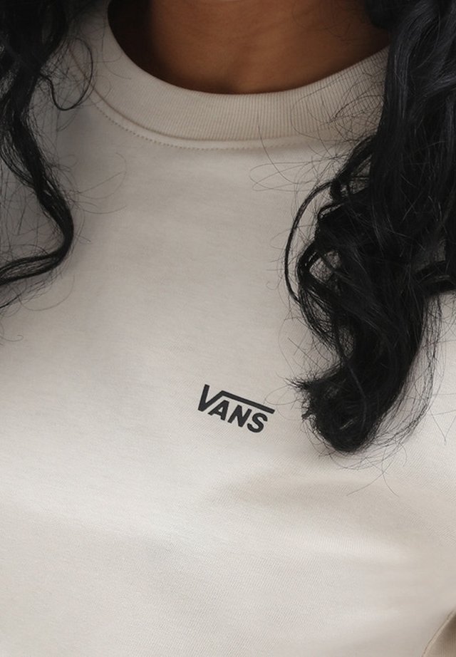 Personne portant un sweat-shirt Vans beige clair avec des cheveux bouclés foncés partiellement visibles autour du col.