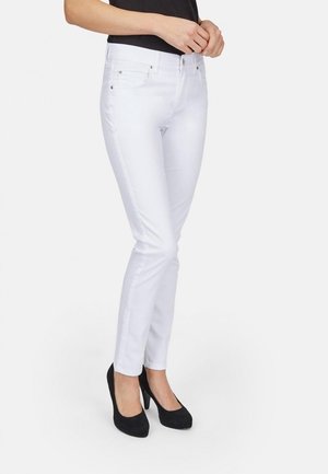 Femme portant un jean blanc ajusté et des talons hauts noirs, debout les mains jointes près de la taille, sur un fond uni.