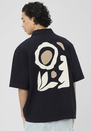 Personne tournée vers la gauche portant un t-shirt noir à manches courtes avec un motif abstrait floral et visage beige et blanc dans le dos.