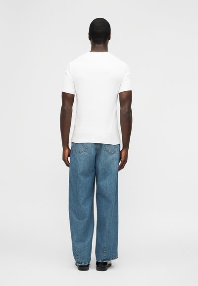 Άνδρας που στέκεται με την πλάτη προς το φακό, φορώντας απλό λευκό κοντομάνικο πουκάμισο, άνετα μπλε τζιν και μαύρα παπούτσια, σε λευκό φόντο.