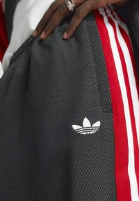 Svarta sportshorts med röda sidoremmar, tillverkade av texturerat tyg. Har den ikoniska vita Adidas-logotypen broderad på benet.