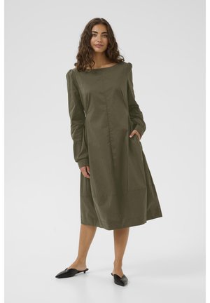 Olive green dress with long sleeves, square neckline, side pockets, and textured cuffs; loose fit with a knee-length hem. Black slip-on shoes.