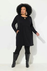 Svart långärmad klänning med nyckelhålsknäppning, texturerat tyg, åtsittande midja och sidofickor, tillsammans med svarta cowboyboots.