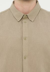 Μπεζ πουκάμισο από ύφασμα με κλασικό γιακά και τέσσερα μπεζ κουμπιά στο εμπρόσθιο μέρος, που φοριέται από ένα άτομο που εμφανίζεται από τον λαιμό μέχρι το στήθος.