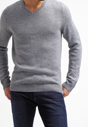 Pull en maille grise avec un col en V, des poignets et un ourlet côtelés, doté d'une texture lisse et d'une coupe ajustée. Porté sur un jean bleu foncé.