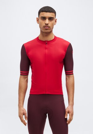 Mlad moški, oblečen v oprijeto rdečo in temno bordo kolesarsko majico s kratkimi rokavi ter ujemajoče se kolesarske kratke hlače, stoji pred enostavnim ozadjem.