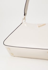 Hvid taske i imiteret læder med trekantet gulddetalje, kontrasterende sort kant, rundede kanter og guldnitter ved bunden.